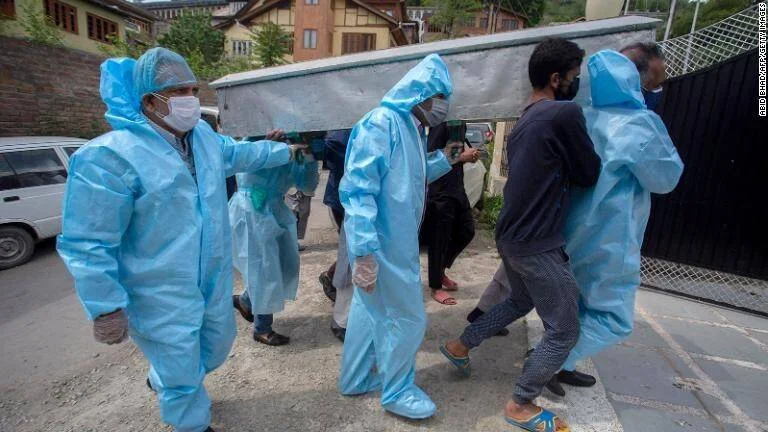 Personas con trajes de protección azules y mascarillas cargan un ataúd junto a familiares durante la pandemia de COVID-19.