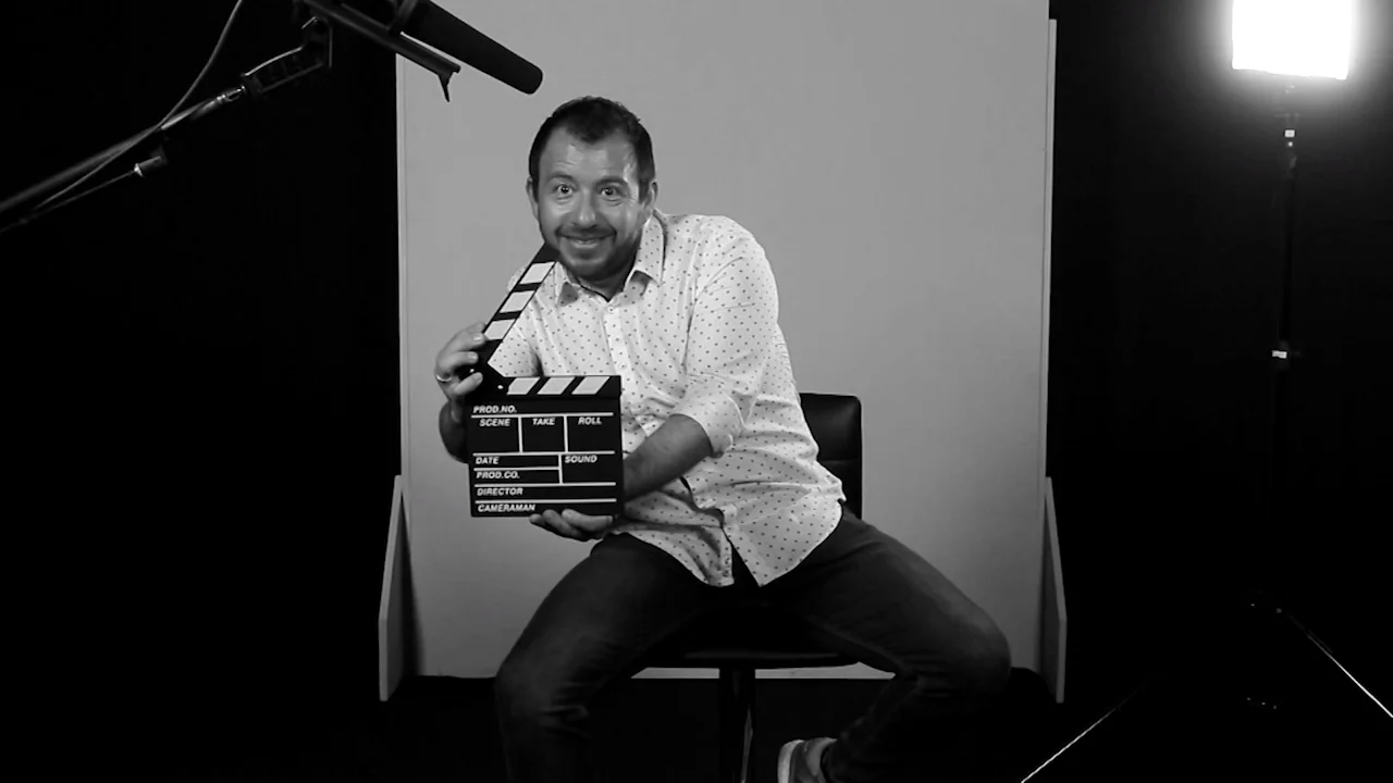 Hombre sonriente sosteniendo una claqueta de filmación mientras posa sentado en un estudio de grabación, en blanco y negro.