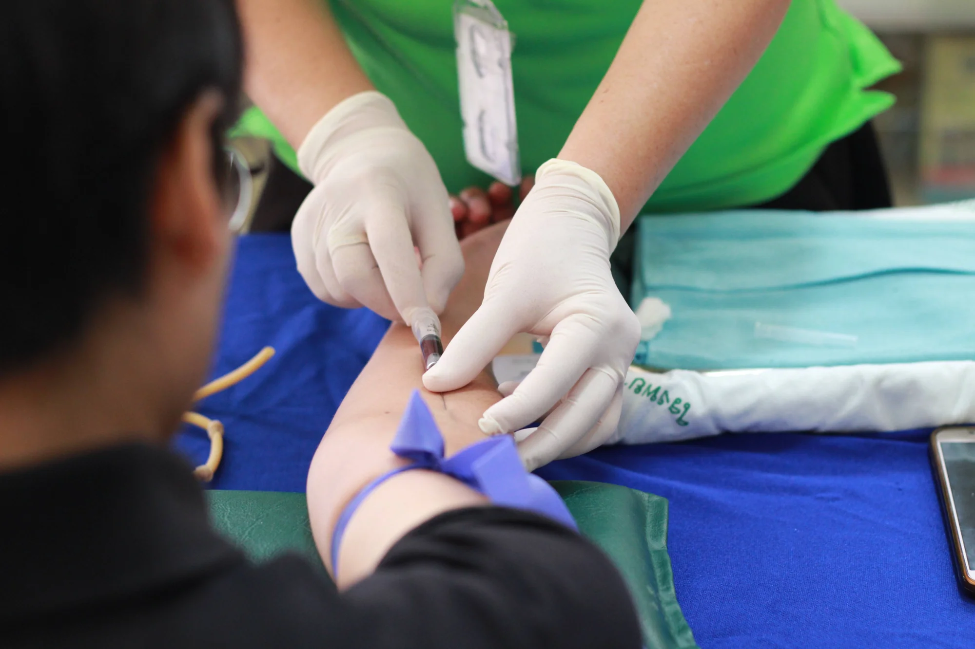 Profesional de salud con guantes extrae sangre del brazo de un paciente con torniquete azul.