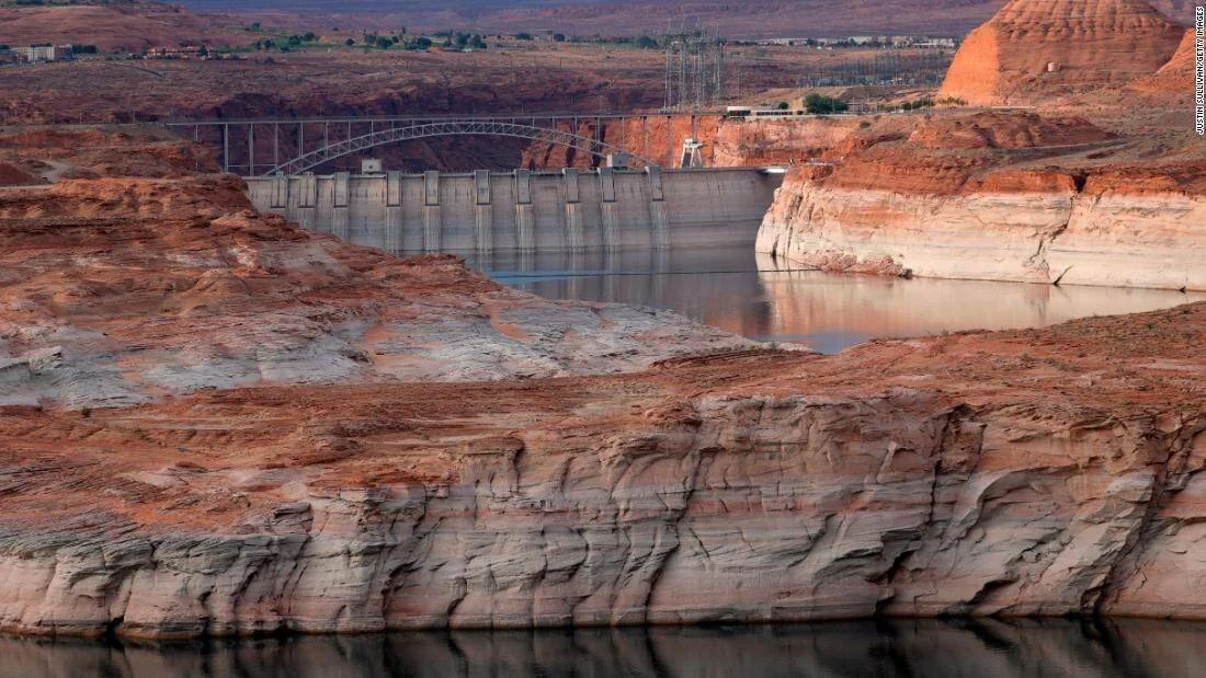 Embalse con nivel de agua críticamente bajo, mostrando una amplia línea de sedimentos blancos en las rocas rojizas junto a una presa.