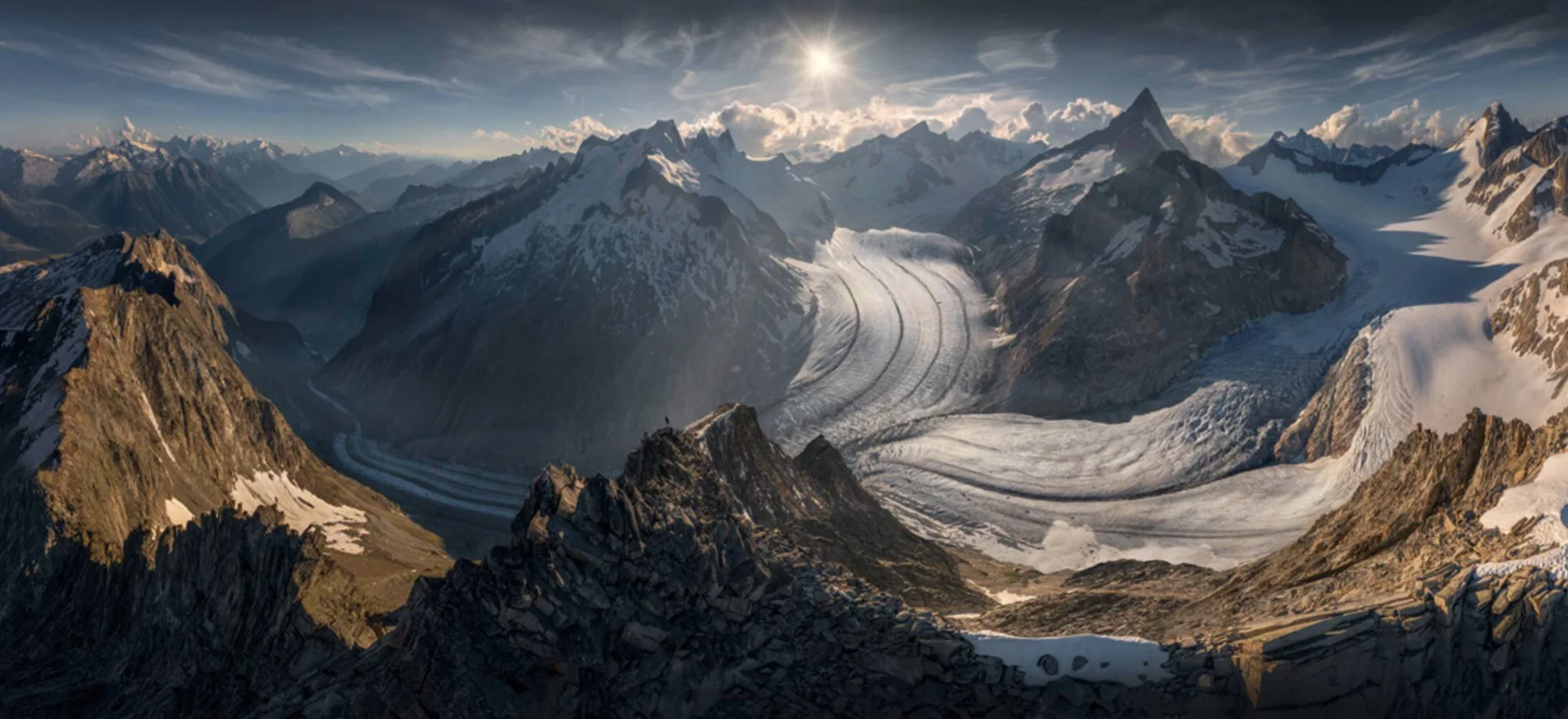 Vista panorámica de un extenso glaciar que desciende entre picos rocosos nevados bajo un cielo con sol brillante.