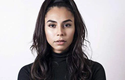 Mujer joven de cabello largo oscuro con remera de cuello alto negra, mirando a cámara con expresión seria.