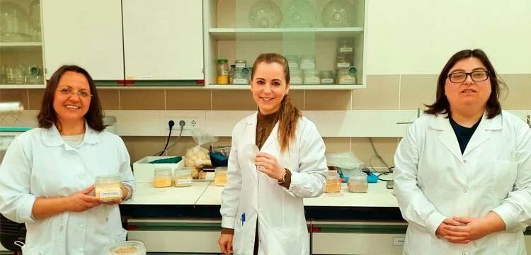 Tres mujeres con batas de laboratorio posan frente a una mesa con frascos de muestras en un laboratorio científico.
