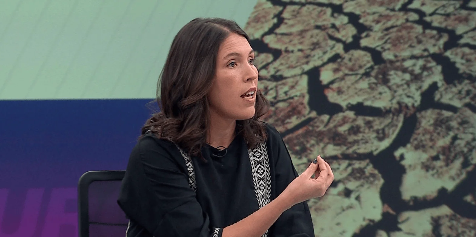 Mujer de cabello oscuro habla gesticulando en un estudio de televisión con pantalla de fondo sobre medio ambiente.