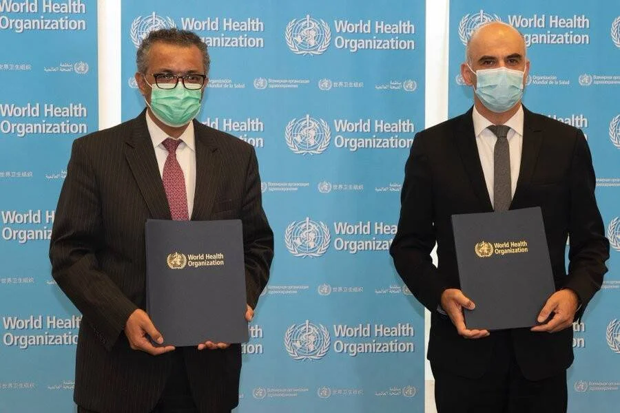 Dos hombres con mascarilla sostienen carpetas de la Organización Mundial de la Salud frente al logo institucional de la OMS.