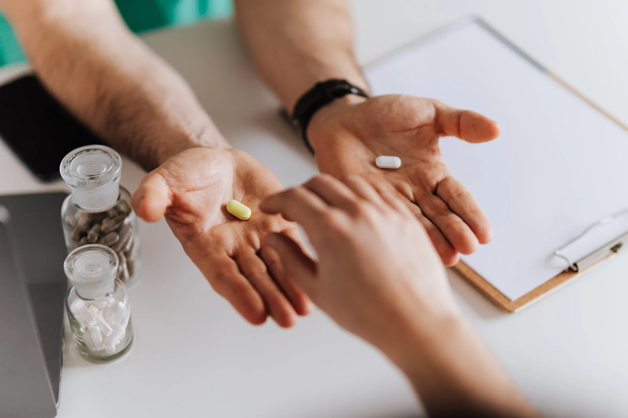 Profesional de salud muestra dos pastillas de diferente color en sus manos abiertas frente a un paciente, junto a frascos de medicamentos.