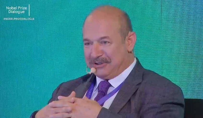Hombre con micrófono de solapa habla durante el Nobel Prize Dialogue, con traje gris y corbata morada.