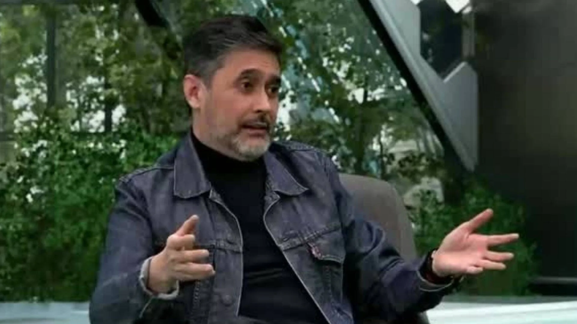 Hombre de mediana edad con campera de jean y cuello alto negro gesticula con ambas manos mientras habla en un estudio de televisión.