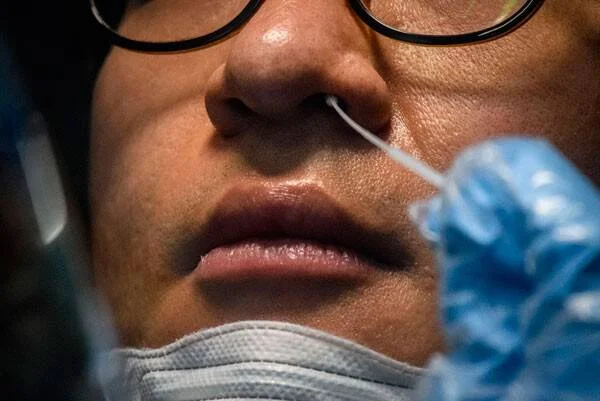 Persona con lentes recibe un hisopo nasal para realizarse un test PCR, con barbijo bajado.