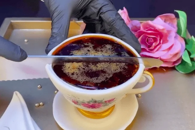 Manos con guantes negros sostienen una cucharilla sobre una taza decorativa llena de líquido oscuro con espuma, junto a una rosa rosa artificial.