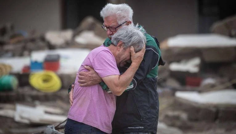 Dos personas mayores se abrazan llorando rodeadas de escombros tras una catástrofe.