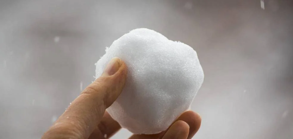 Una mano sostiene una bola de nieve compacta con copos cayendo al fondo.