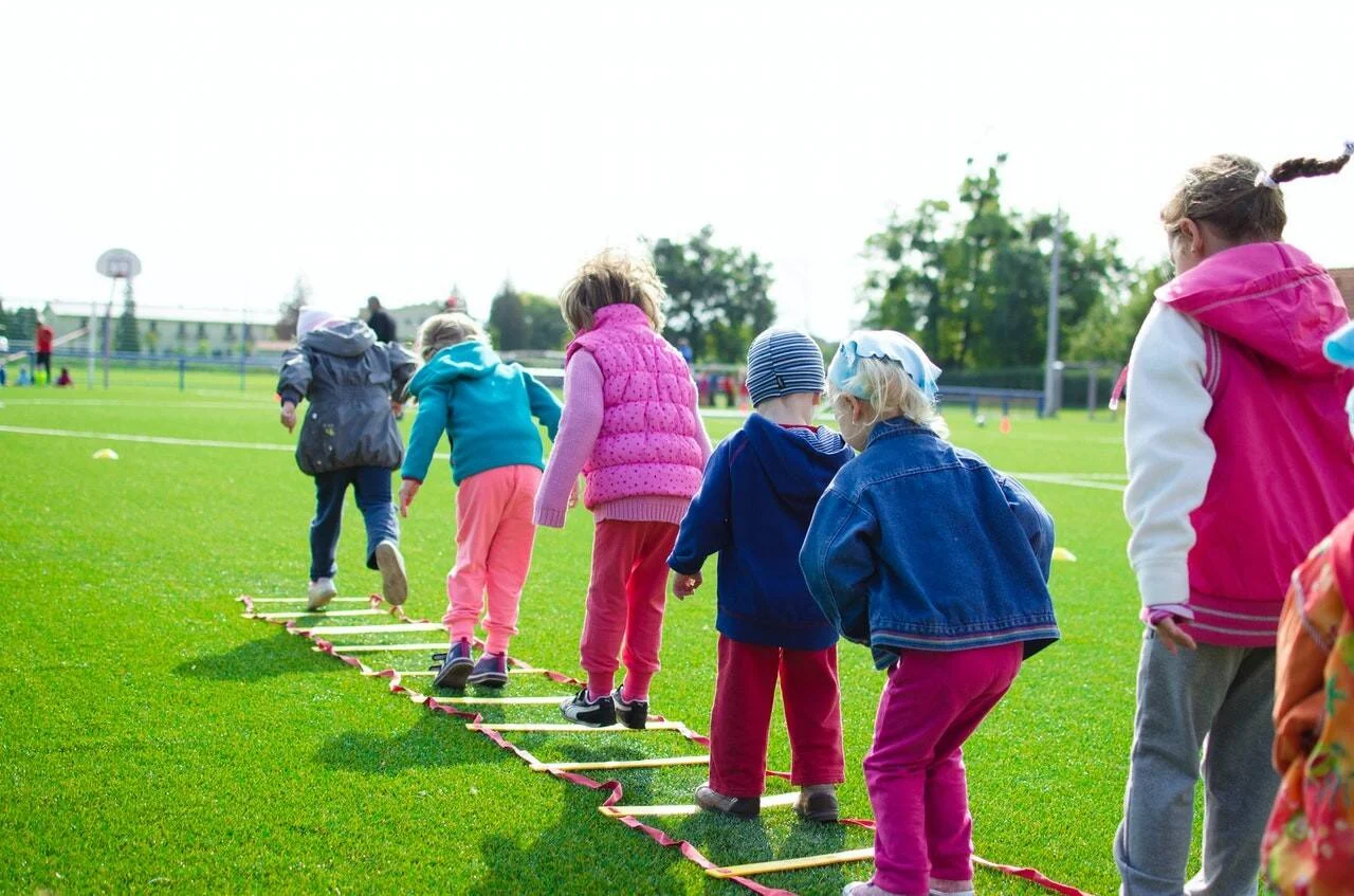 Grupo de niños pequeños realiza ejercicios de coordinación con una escalera de agilidad en un campo deportivo al aire libre.