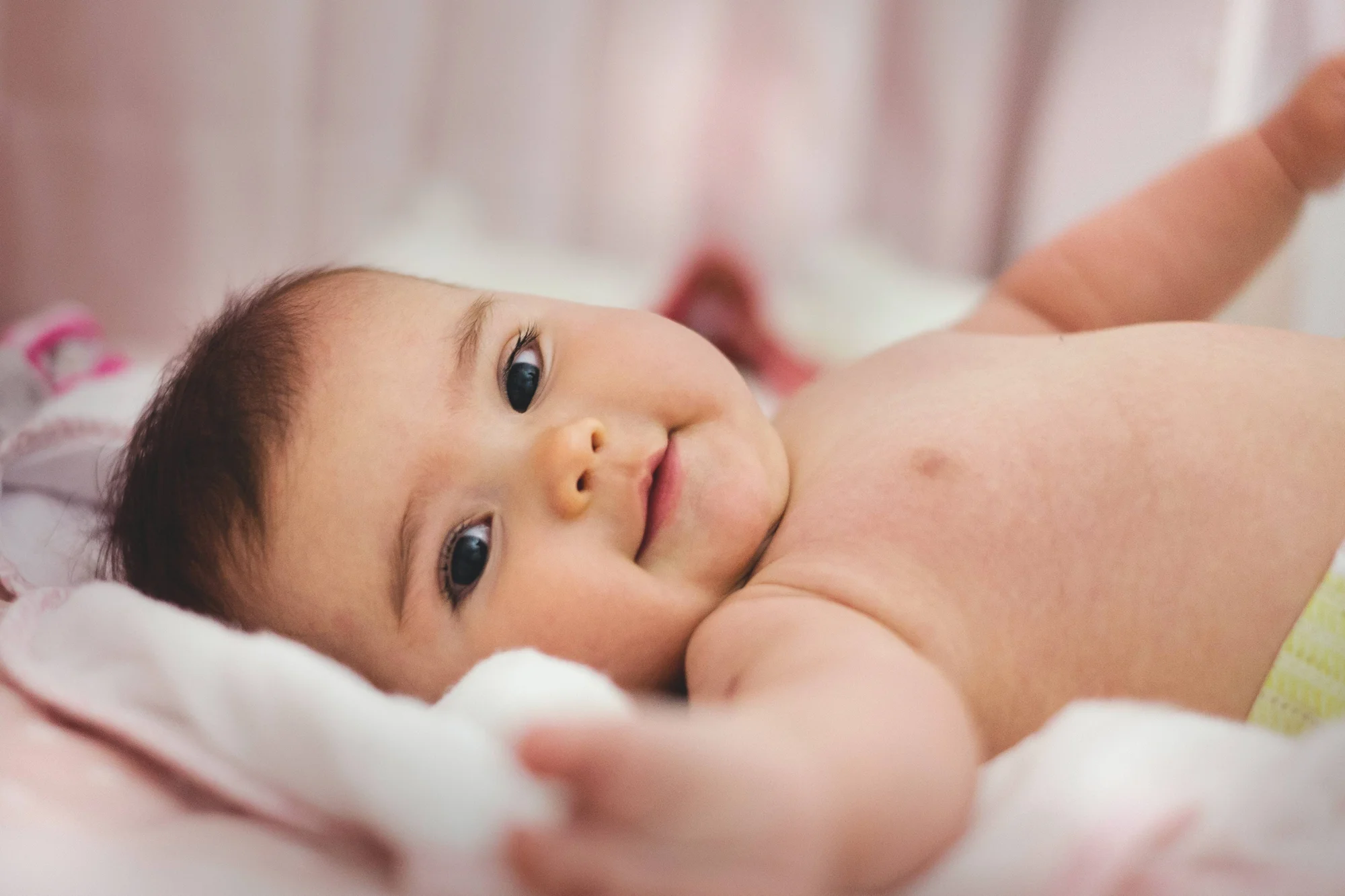 Bebé de pocos meses acostado boca arriba, con los ojos abiertos y una leve sonrisa, sobre una superficie blanda rosada.