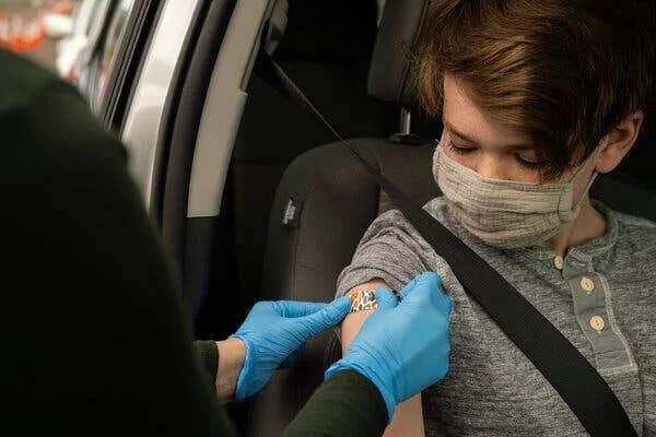 Un niño con mascarilla recibe una vacuna en su brazo dentro de un vehículo, mientras un profesional de salud con guantes azules coloca una curita.