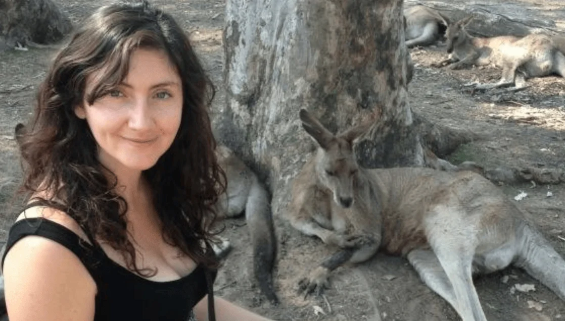 Mujer de cabello oscuro sonríe junto a varios canguros descansando bajo un árbol en un entorno natural.