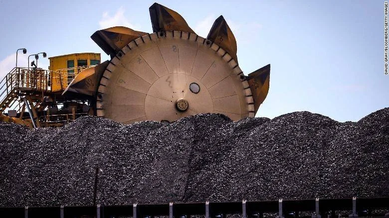 Gran rueda de excavadora minera amarilla operando sobre una enorme pila de carbón en un puerto o planta industrial.