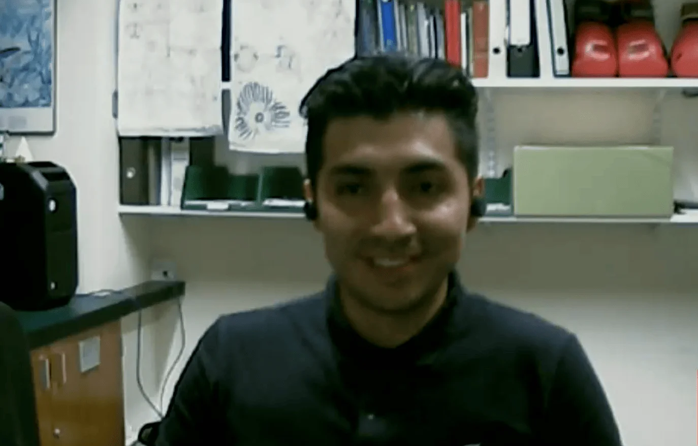 Joven hombre sonriente frente a estantes con carpetas y guantes de boxeo rojos, en una videollamada desde una oficina.