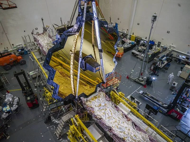 El espejo primario hexagonal dorado de un telescopio espacial es izando en una sala limpia durante su ensamblaje.