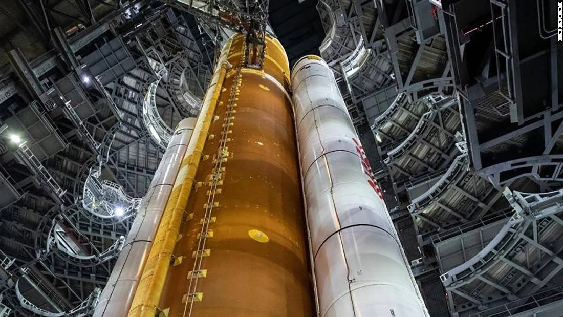 Vista desde abajo del cohete SLS de la NASA con sus propulsores laterales blancos y el núcleo naranja dentro del edificio de ensamblaje.