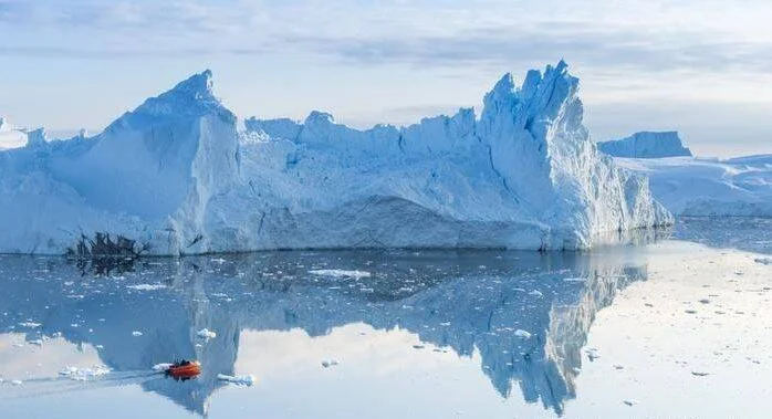 Una pequeña embarcación naranja navega entre bloques de hielo frente a enormes icebergs que se reflejan en aguas calmas del Ártico o Antártida.