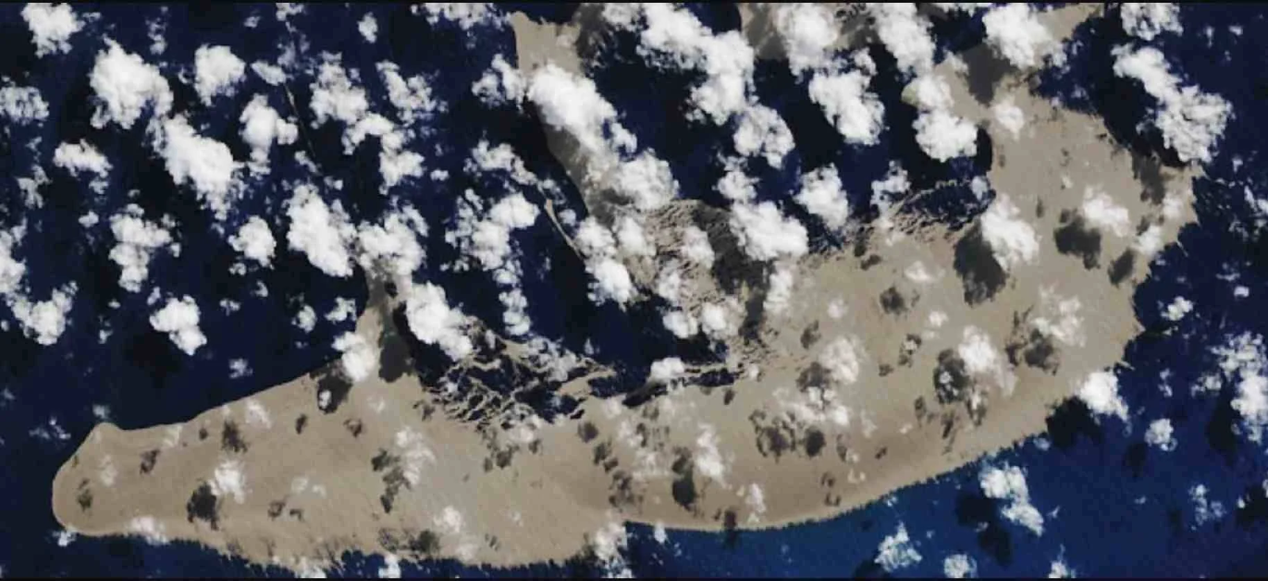 Vista satelital de una isla de origen volcánico de color grisáceo rodeada de océano azul oscuro y nubes blancas dispersas.