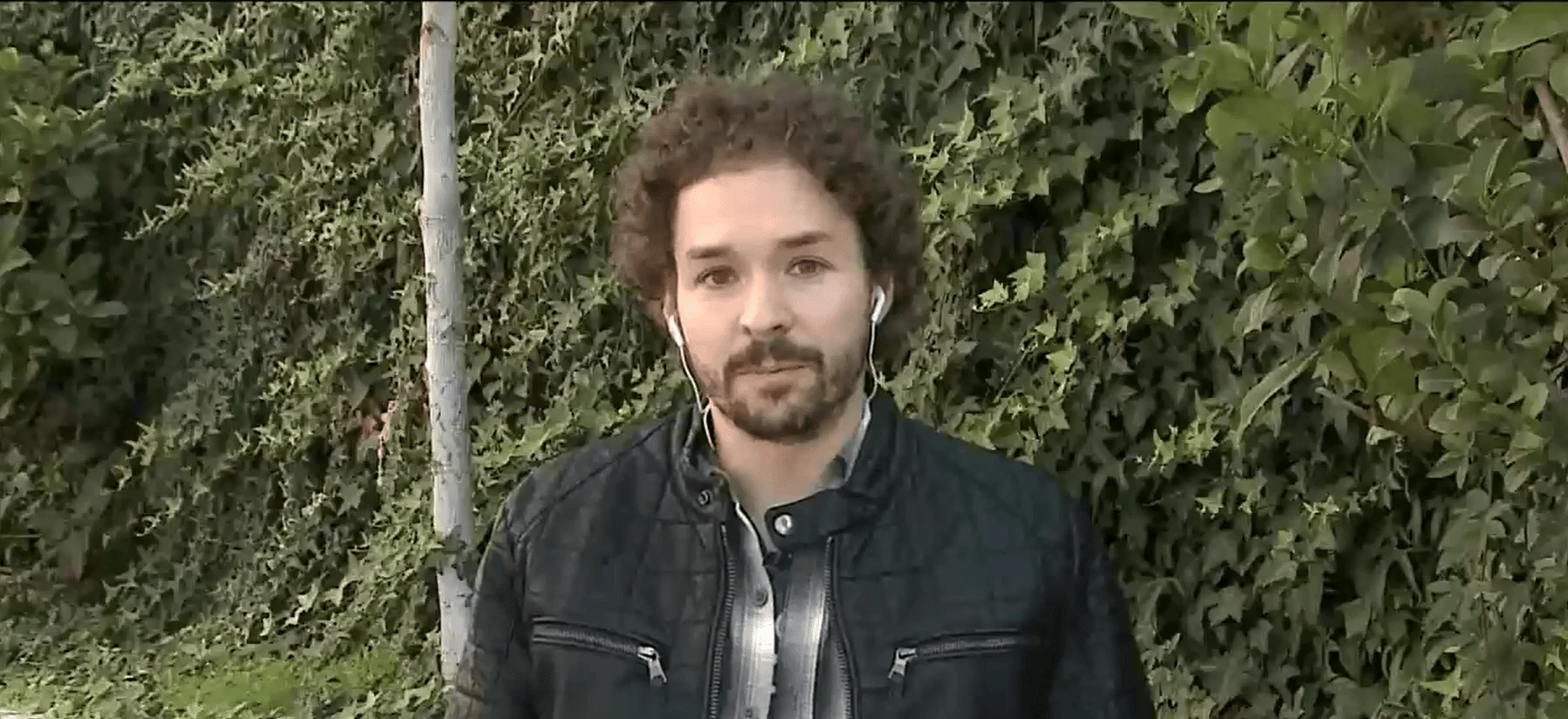 Hombre joven de cabello rizado y barba, con auriculares y chaqueta negra, frente a una pared cubierta de hiedra.