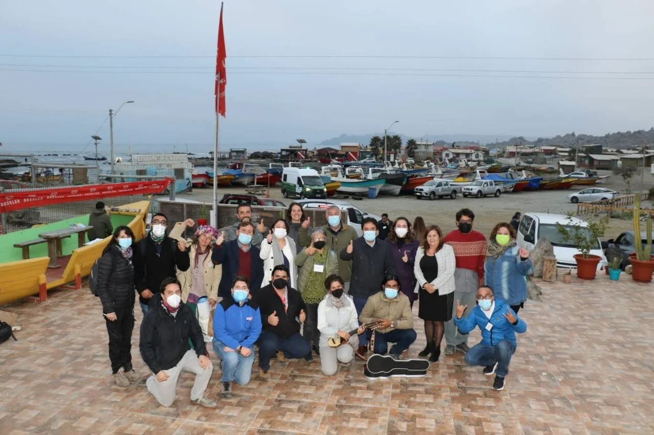 Grupo de aproximadamente veinte personas con mascarillas posan con pulgares arriba frente a una caleta pesquera con botes coloridos al fondo.