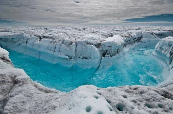 Canal de agua turquesa fluyendo sobre la superficie de un glaciar bajo un cielo nublado.