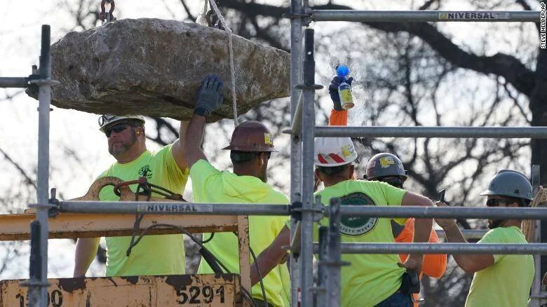 Trabajadores con cascos y chalecos amarillos guían un gran bloque de piedra suspendido por una grúa sobre un andamio.