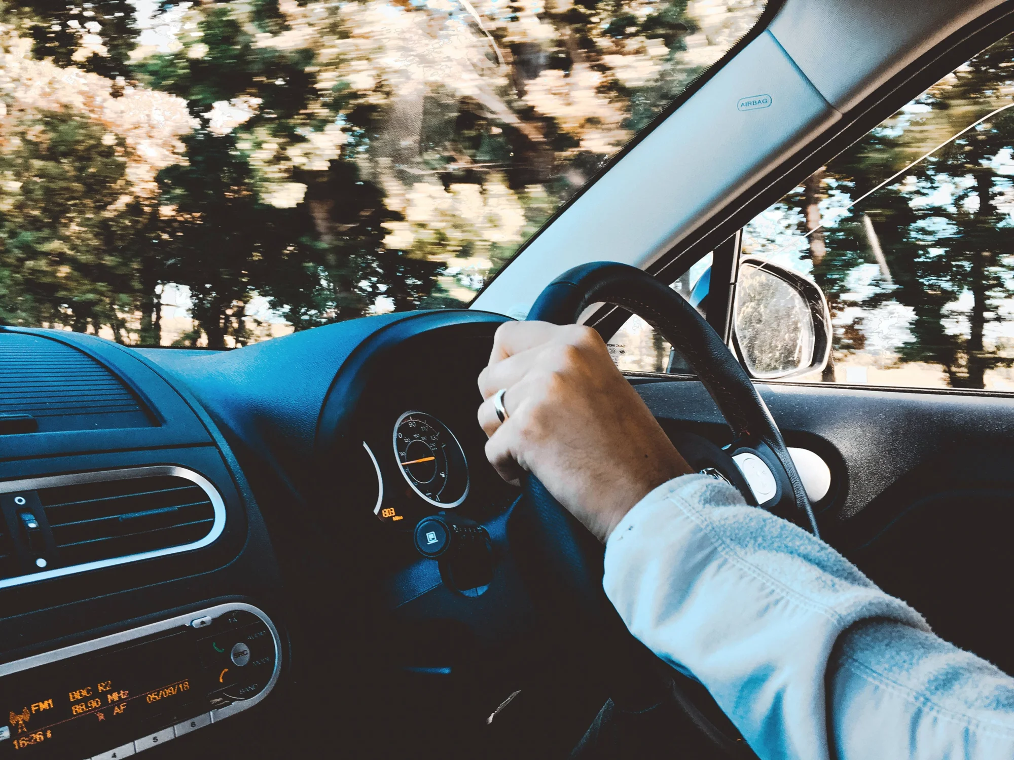 Conductor sujeta el volante de un automóvil mientras circula por una carretera rodeada de árboles.