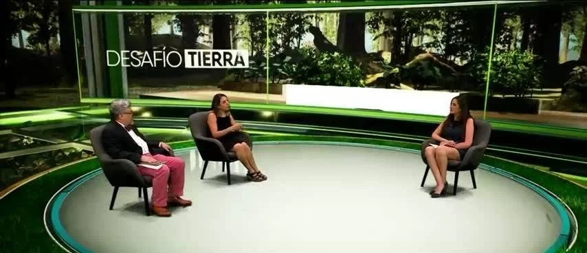 Tres personas conversan sentadas en sillas en el estudio del programa 'Desafío Tierra', con escenografía verde y pantalla con árboles al fondo.