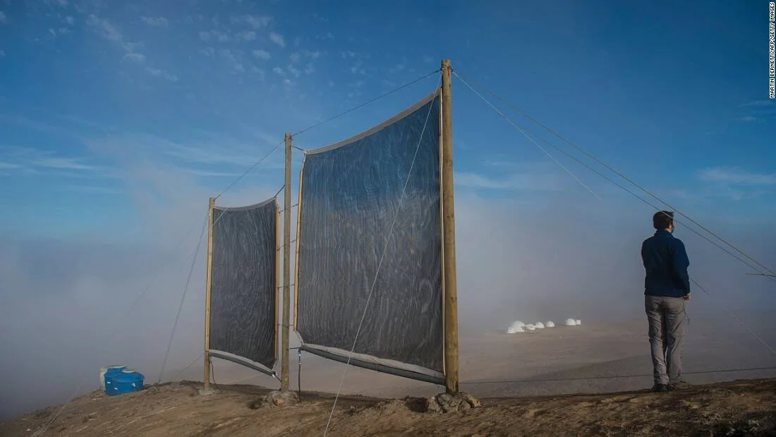 Una persona observa grandes mallas captadoras de niebla instaladas en una colina árida para recolectar agua del ambiente.