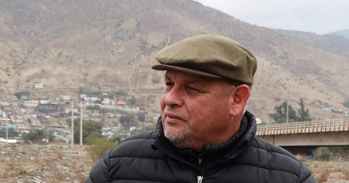 Hombre adulto con gorra verde y chaqueta negra mirando hacia un lado, con un poblado en ladera de cerro al fondo.