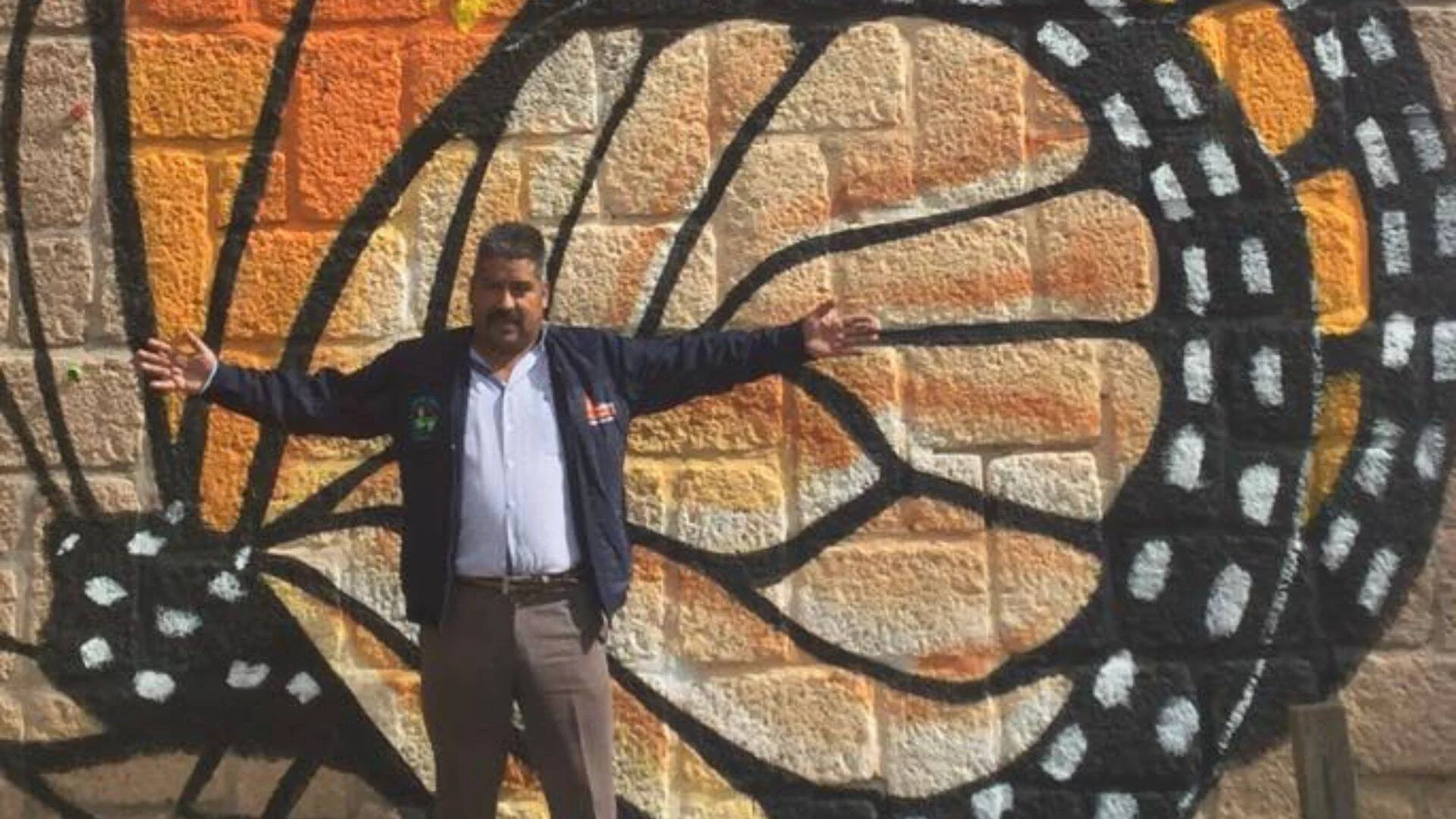 Hombre con los brazos abiertos frente a un mural en piedra con una gran mariposa monarca naranja y negra.