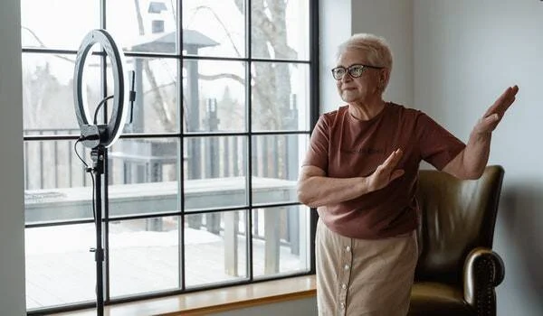 Mujer mayor con gafas realiza ejercicios frente a un aro de luz para grabar contenido en casa.