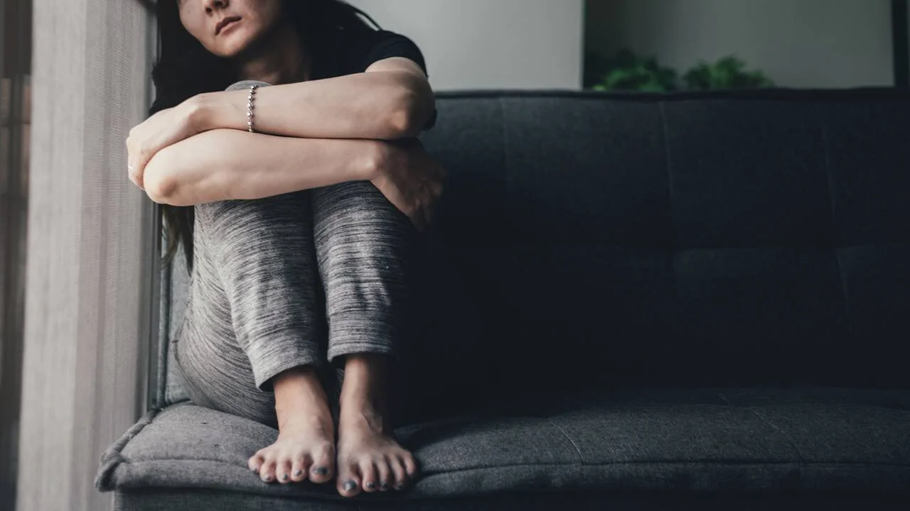 Mujer joven sentada en un sofá con las rodillas abrazadas al pecho, en postura de tristeza o angustia emocional.
