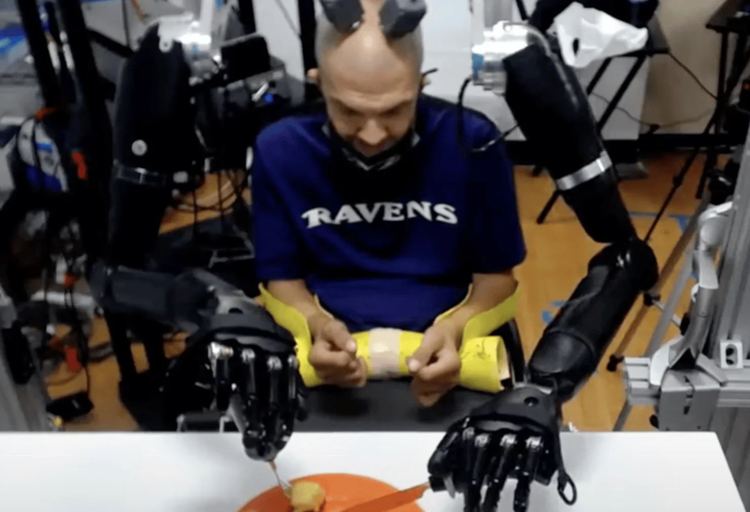 Paciente con electrodos en la cabeza controla dos brazos robóticos mientras manipula un objeto con sus propias manos en un laboratorio.