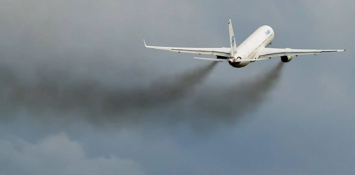 Avión comercial blanco despega dejando una estela de humo negro oscuro sobre un cielo nublado.