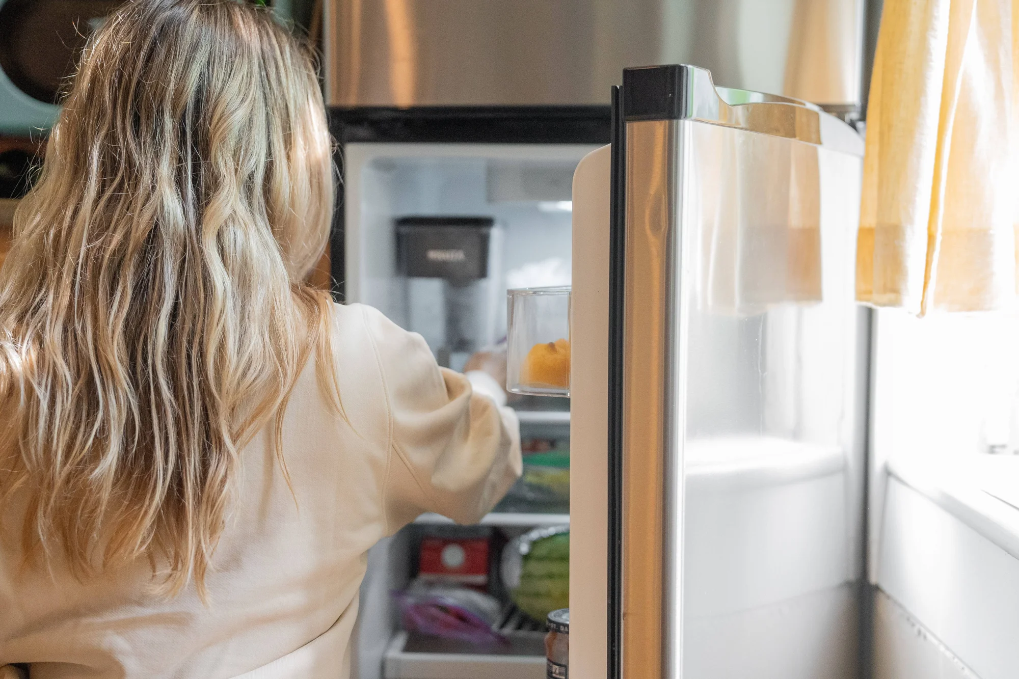 Mujer de espaldas con cabello rubio busca un alimento en el interior de un refrigerador doméstico abierto.