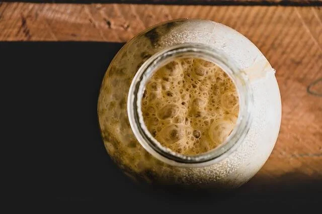 Vista cenital de un frasco de vidrio con líquido marrón burbujeante, posiblemente masa madre o bebida fermentada.