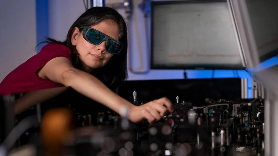 Mujer con gafas de protección láser ajusta componentes de un equipo óptico en un laboratorio con iluminación azul.