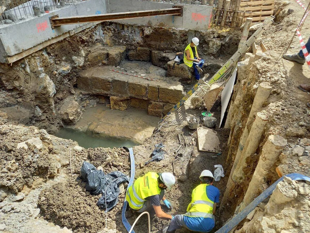 Tres arqueólogos con chalecos reflectantes trabajan en una excavación urbana donde se han descubierto muros de piedra antiguos.