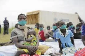 Mujeres y niños con mascarillas quirúrgicas sentados al aire libre en un campo de atención humanitaria.