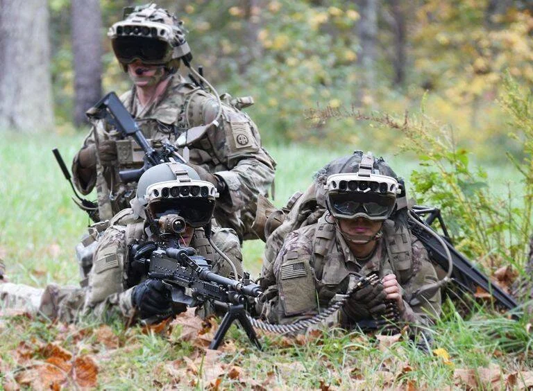 Tres soldados con equipamiento táctico y visores de realidad aumentada en posición de combate durante un ejercicio militar en zona boscosa.