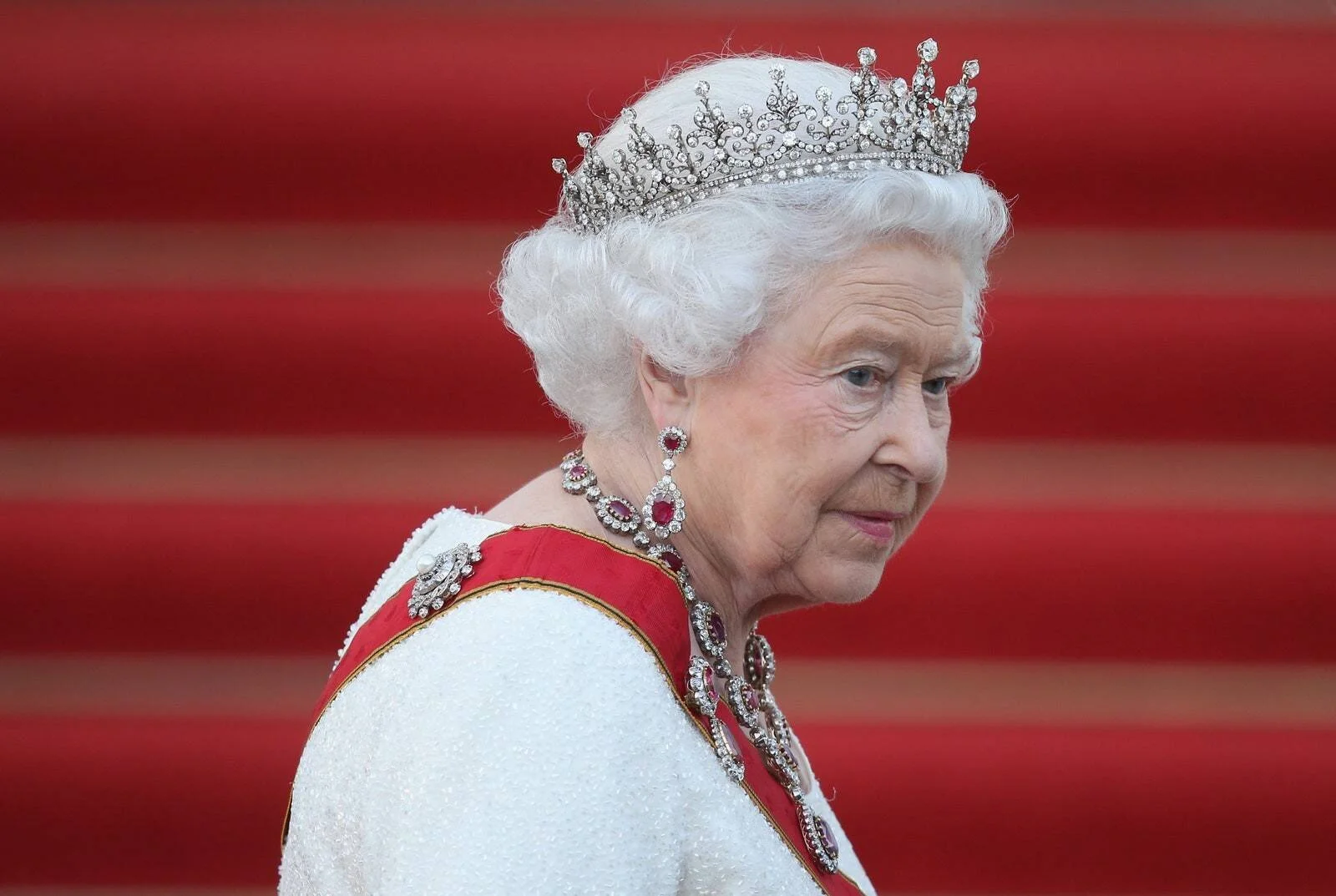 Mujer mayor con cabello blanco, tiara de diamantes, collar y pendientes de rubíes, y banda roja sobre vestido blanco en acto de estado.