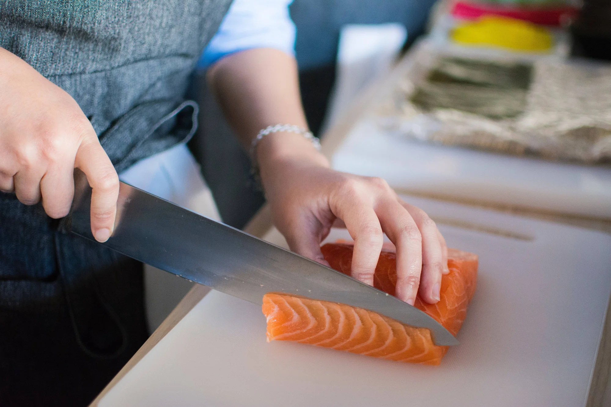 Manos cortando un filete de salmón crudo en láminas finas sobre una tabla blanca, con hojas de nori al fondo.