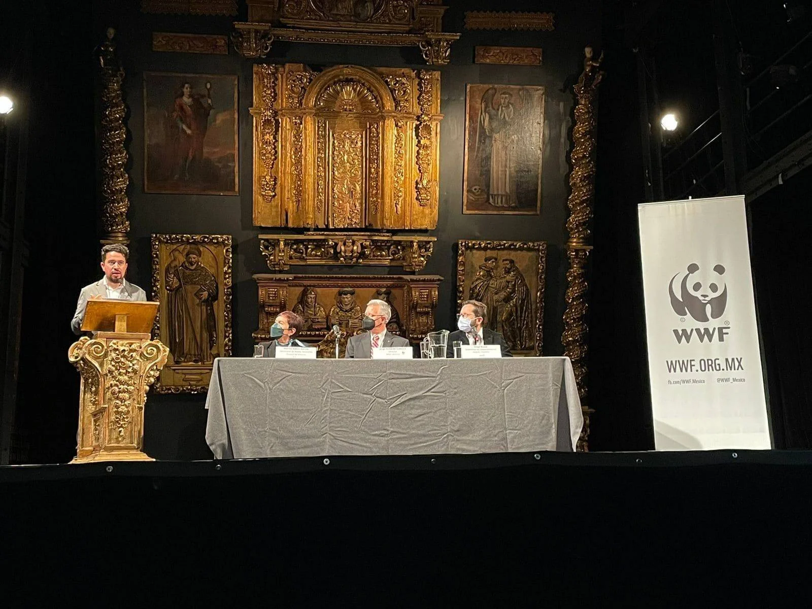 Panel de cuatro personas en conferencia de WWF México, con un orador en el atril y tres panelistas sentados ante retablo dorado colonial.