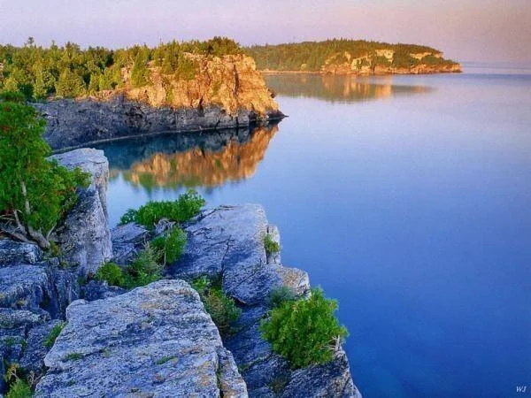 Acantilados de roca caliza con vegetación verde junto a un lago de aguas azules calmas al atardecer.