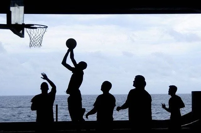Siluetas de cinco personas jugando baloncesto al aire libre con el mar y un cielo nublado de fondo.